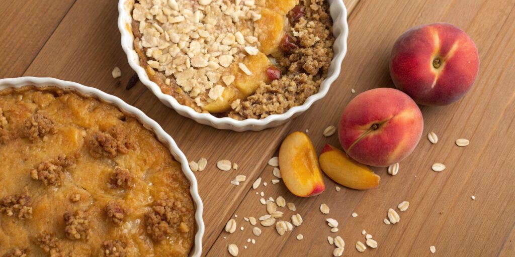 Top-down view comparing a peach crumble with a smooth golden topping and a peach crisp with a crunchy oat-and-nut topping, surrounded by fresh peaches on a wooden countertop